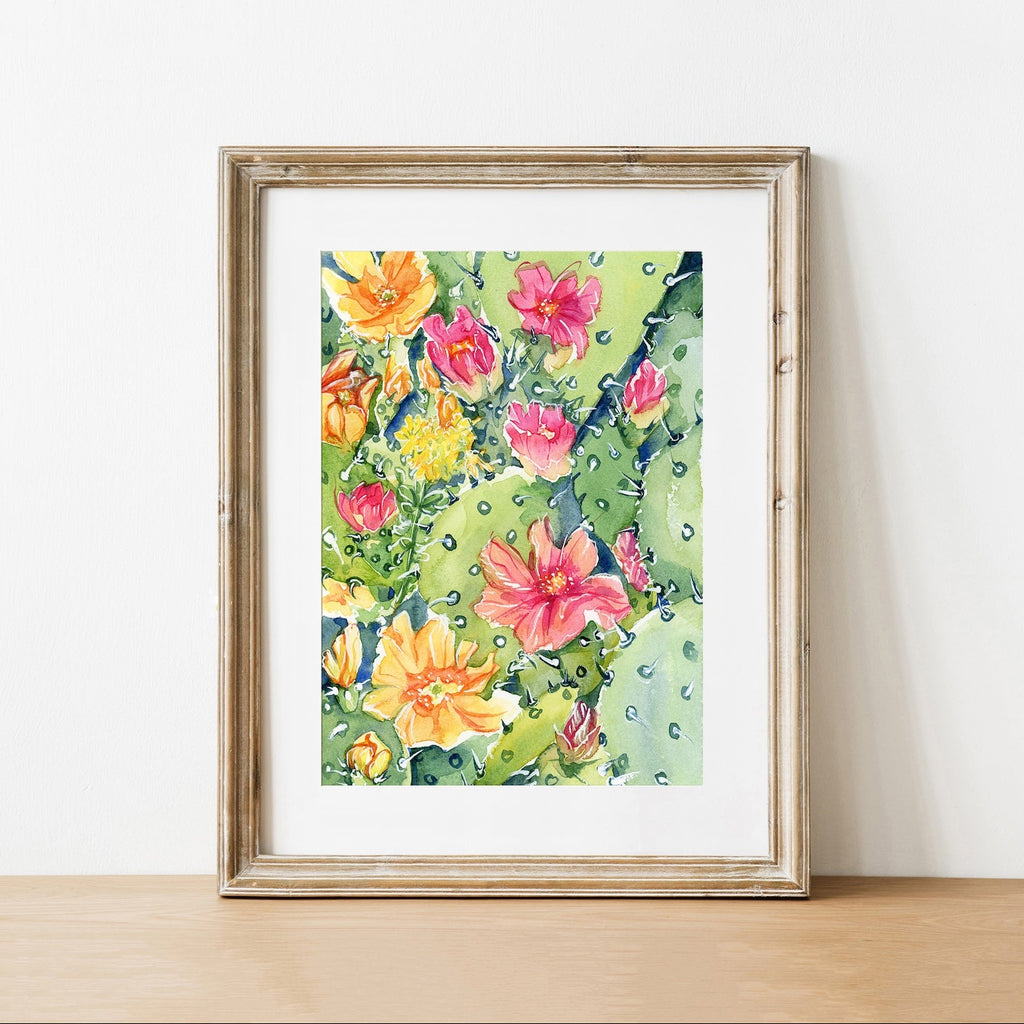 Painting of pink and yellow prickly pear cactus blossoms in a rustic wood frame leaning against a white wall.